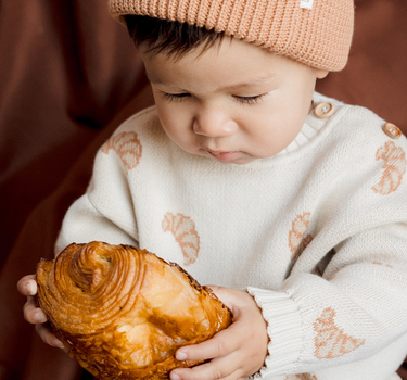 Baby boy looking at croissant while wearing organic cotton knit sweater with croissant jacquard pattern with Magnetic Buttons at shoulder.