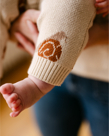 Image shows baby girl wearing organic cotton knit romper with jacquard snail pattern in oatmeal color.