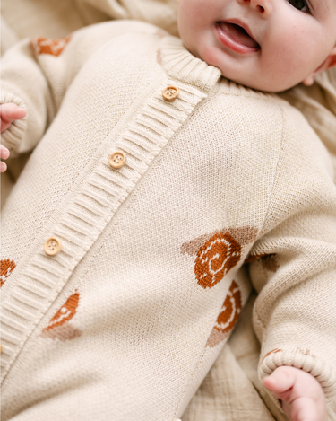 Image shows baby girl wearing organic cotton knit romper with jacquard snail pattern in oatmeal color.