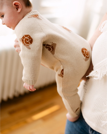 Image shows baby girl wearing organic cotton knit romper with jacquard snail pattern in oatmeal color.