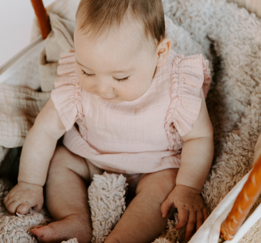 Image shows baby girl wearing organic cotton muslin flutter sleeve romper in orchid pink with magnetic closures at the crotch.