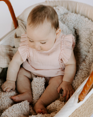 Image shows baby girl wearing organic cotton muslin flutter sleeve romper in orchid pink with magnetic closures at the crotch.