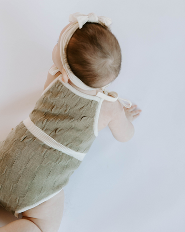 Baby girl wearing organic cotton knit bubble romper in truffle with tie shoulder straps and magnetic closure at crotch.