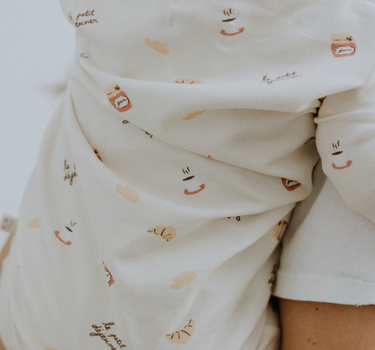 Baby wearing modal pajama set in Petit Déjeuner print with coffee mugs, croissants, butter, and jam.