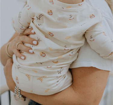 Baby wearing modal pajama set in Petit Déjeuner print with coffee mugs, croissants, butter, and jam.