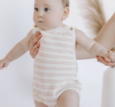 Image shows baby girl wearing organic cotton knit stripe bubble romper in pink stripes with adjustable shoulder straps and magnetic closures at the crotch for easy changing.