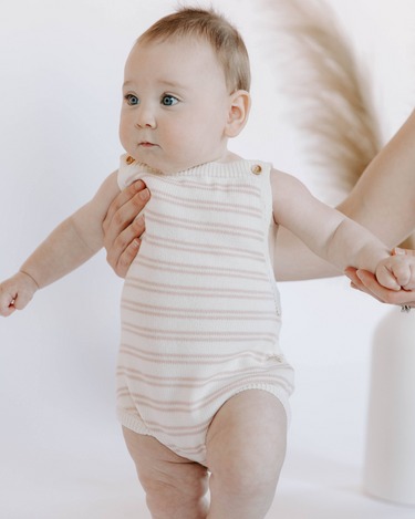 Image shows baby girl wearing organic cotton knit stripe bubble romper in pink stripes with adjustable shoulder straps and magnetic closures at the crotch for easy changing.