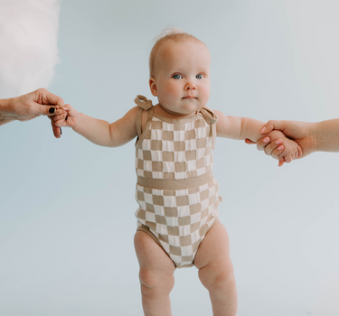 Picture of baby girl wearing organic cotton knit bubble romper in beige checkers with tie shoulder strings and hidden magnetic closures at crotch for easy changing.