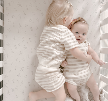 Image shows siblings cuddling in crib wearing organic cotton stripe knit set in mushroom and stripe romper in mushroom. 