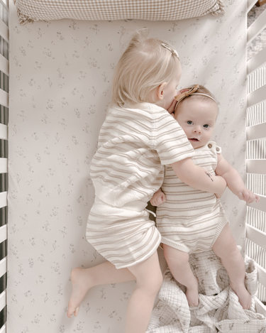 Image shows siblings cuddling in crib wearing organic cotton stripe knit set in mushroom and stripe romper in mushroom. 