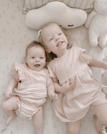 Little girl laying on bed wearing organic cotton muslin flutter sleeve dress in orchid pink.