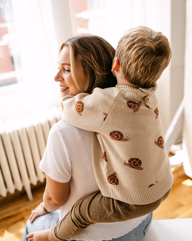 Image shows toddler boy wearing organic cotton knit sweater with snail jacquard pattern in oatmeal.