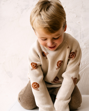 Image shows toddler boy wearing organic cotton knit sweater with snail jacquard pattern in oatmeal.