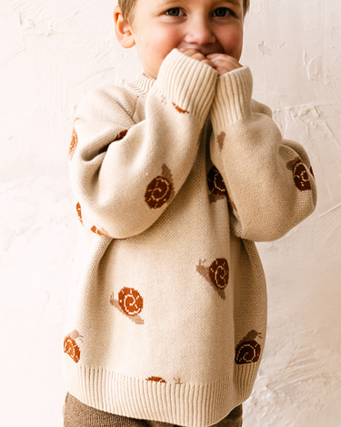 Image shows toddler boy wearing organic cotton knit sweater with snail jacquard pattern in oatmeal.