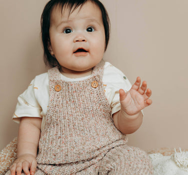 Baby girl wearing 100% organic cotton knit overalls in rose melange color with adjustable wood buttons.