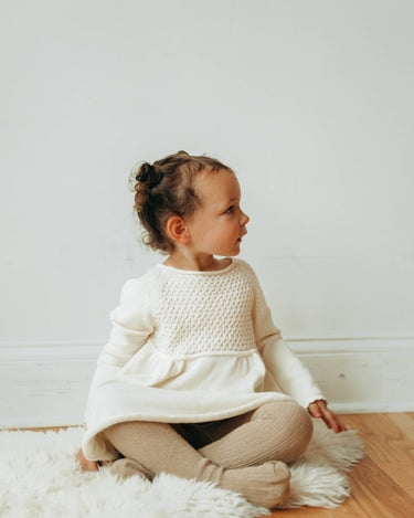 Image of little girl wearing knit dress in oatmilk color with sweet details and button detail at the back of the neck.