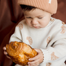 Baby boy looking at croissant while wearing organic cotton knit sweater with croissant jacquard pattern with Magnetic Buttons at shoulder.