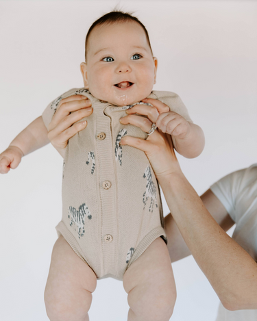 Image shows baby girl wearing organic cotton knit playsuit with zebra jacquard and magnetic button closures in mushroom beige color.