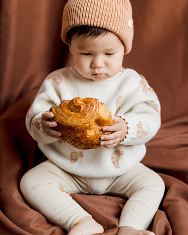 Image shows baby boy wearing organic cotton knit croissant sweater and organic cotton knit beanie in acorn.