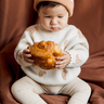 Image shows baby boy wearing organic cotton knit croissant sweater and organic cotton knit beanie in acorn.