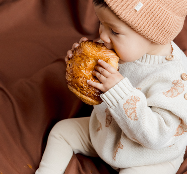 Image shows baby boy wearing organic cotton knit croissant cardigan and organic cotton knit beanie in acorn.