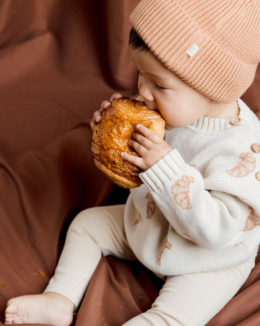 Image shows baby boy wearing organic cotton knit croissant cardigan and organic cotton knit beanie in acorn.