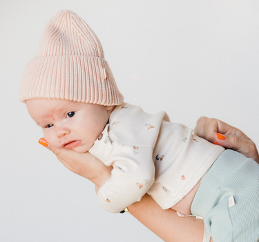 Picture shows baby gear in mother's arms wearing organic cotton rib knit beanie in raspberry macaron color. 