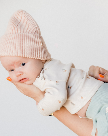 Picture shows baby gear in mother's arms wearing organic cotton rib knit beanie in raspberry macaron color. 