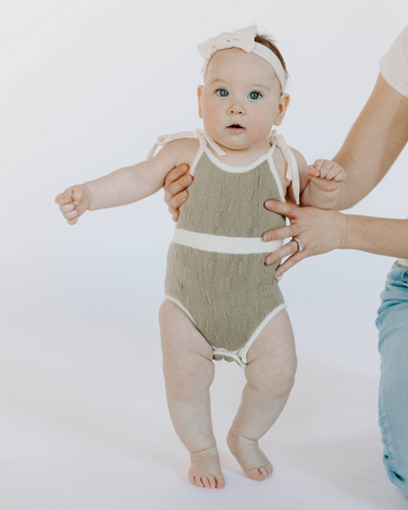 Baby girl wearing organic cotton knit bubble romper in truffle with magnetic opening at crotch and tie shoulder straps.