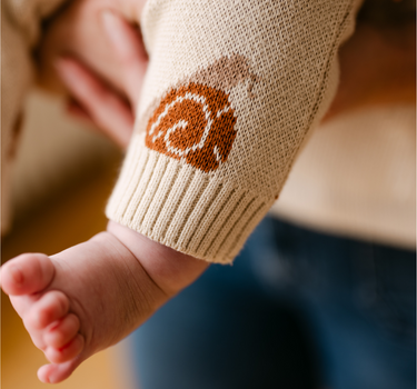 Image shows baby girl wearing organic cotton knit romper with jacquard snail pattern in oatmeal color. 