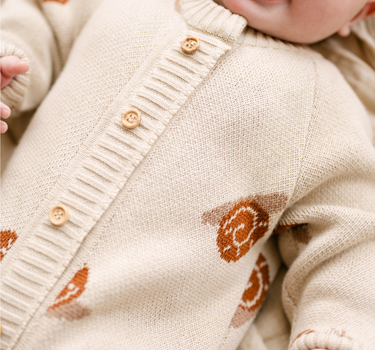 Image shows baby girl wearing organic cotton knit romper with jacquard snail pattern in oatmeal color. 