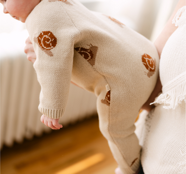 Image shows baby girl wearing organic cotton knit romper with jacquard snail pattern in oatmeal color. 