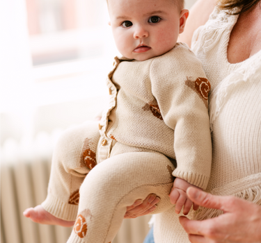 Image shows baby girl wearing organic cotton knit romper with jacquard snail pattern in oatmeal color. 