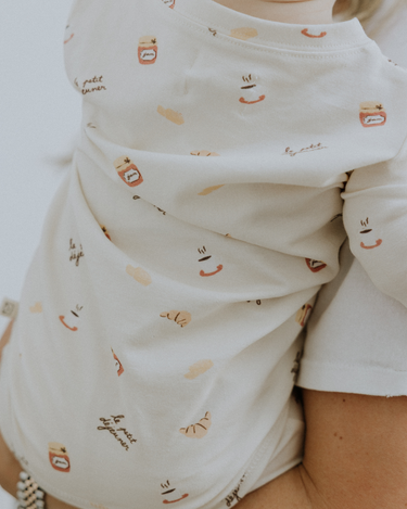 Baby wearing modal convertible footy pajamas in Petit Déjeuner print with coffee mugs, croissants, butter, and jam.