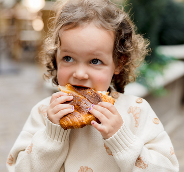 Picture shows toddler girl eating a croissant while wearing an organic cotton knit sweater with croissant jacquard pattern and Magnetic Buttons at the shoulder. 