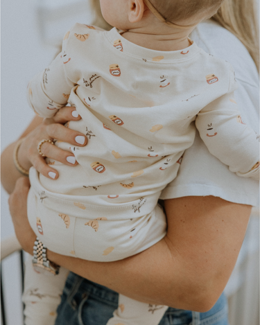 Baby wearing modal pajama set in Petit Déjeuner print with coffee mugs, croissants, butter, and jam.