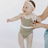 Baby girl wearing organic cotton knit bubble romper in truffle with tie shoulder straps and magnetic closure at crotch.