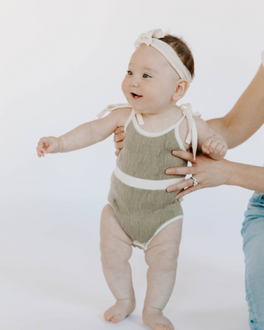 Baby girl wearing organic cotton knit bubble romper in truffle with tie shoulder straps and magnetic closure at crotch.
