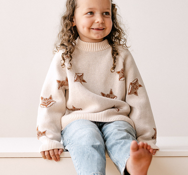 Image shows little girl wearing organic cotton knit sweater with jacquard fox pattern in pale pink. 