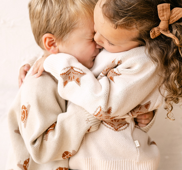 Image shows little girl wearing organic cotton knit sweater with jacquard fox pattern in pale pink and little boy wearing organic cotton knit sweater with jacquard snail pattern in oatmeal. 