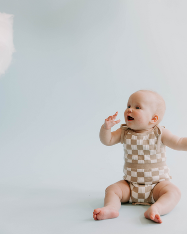 Baby girl wearing organic cotton bubble romper with hidden, sewn-in magnets at crotch and adjust tie strings at shoulders in beige checkers.