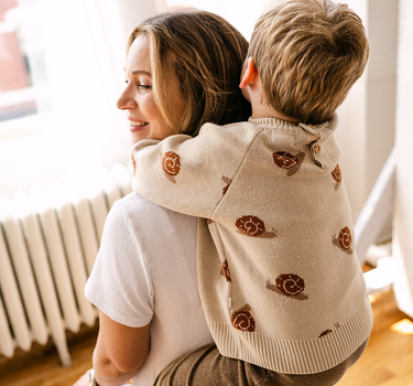 Image shows toddler boy wearing organic cotton knit sweater with snail jacquard pattern in oatmeal. 