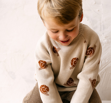 Image shows toddler boy wearing organic cotton knit sweater with snail jacquard pattern in oatmeal. 