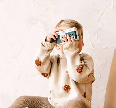 Image shows toddler boy wearing organic cotton knit sweater with snail jacquard pattern in oatmeal. 