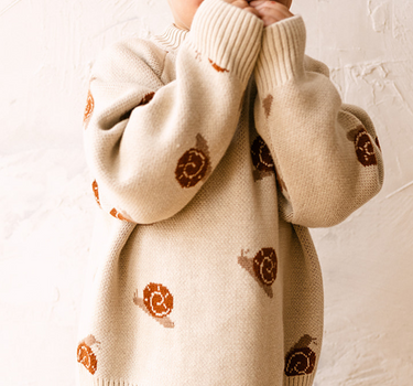 Image shows toddler boy wearing organic cotton knit sweater with snail jacquard pattern in oatmeal. 