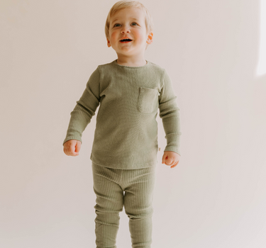 Image of little boy wearing organic cotton rib two piece set in neutral basil color. Set includes a long sleeve top with Magnetic Buttons at the neck in sizes through 12-18 months as well as leggings with a faux drawstring.