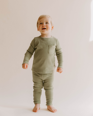 Image of little boy wearing organic cotton rib two piece set in neutral basil color. Set includes a long sleeve top with Magnetic Buttons at the neck in sizes through 12-18 months as well as leggings with a faux drawstring.